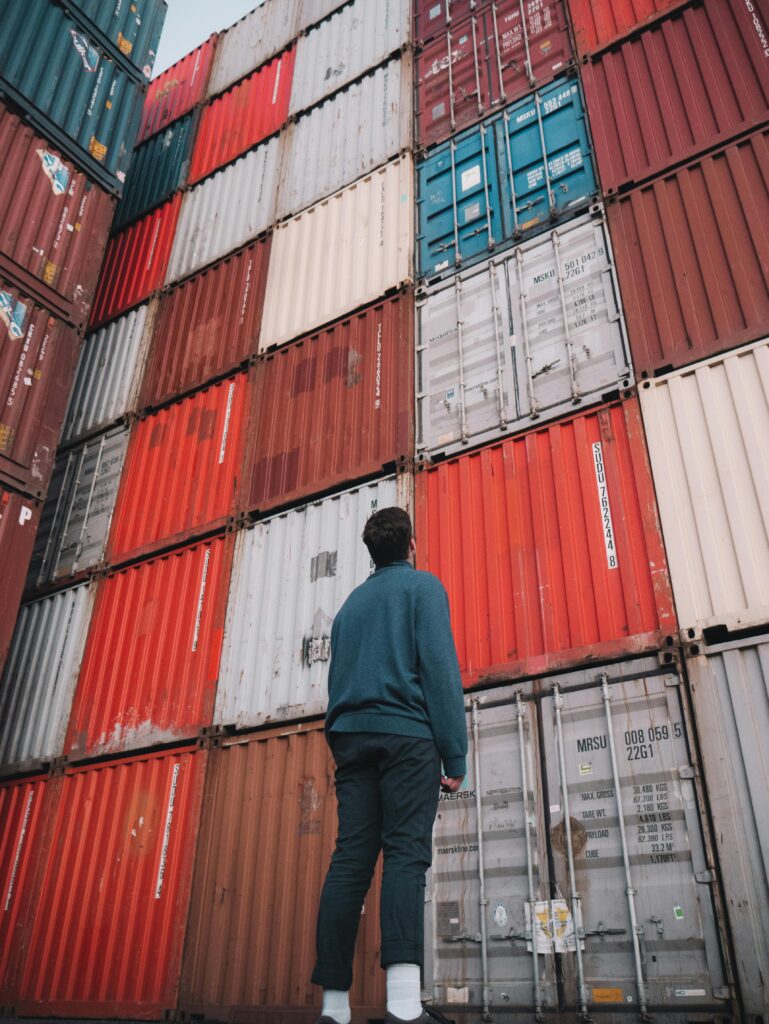 A man stands looking up at a stack of colorful cargo containers at a port, capturing the essence of global trade and commerce.
